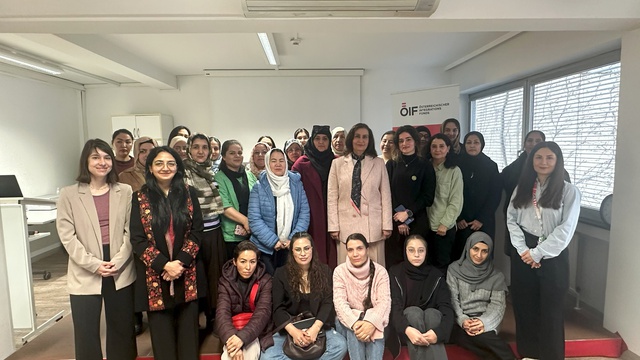 Gruppenfoto von etwa 30 Frauen in einem Raum. Die Teilnehmerinnen stehen auf einer roten Stufe und sind in unterschiedlichen Outfits gekleidet. Der Hintergrund zeigt Fenster und eine Präsentationswand mit einem Logo. Sie wirken konzentriert und freundlich.