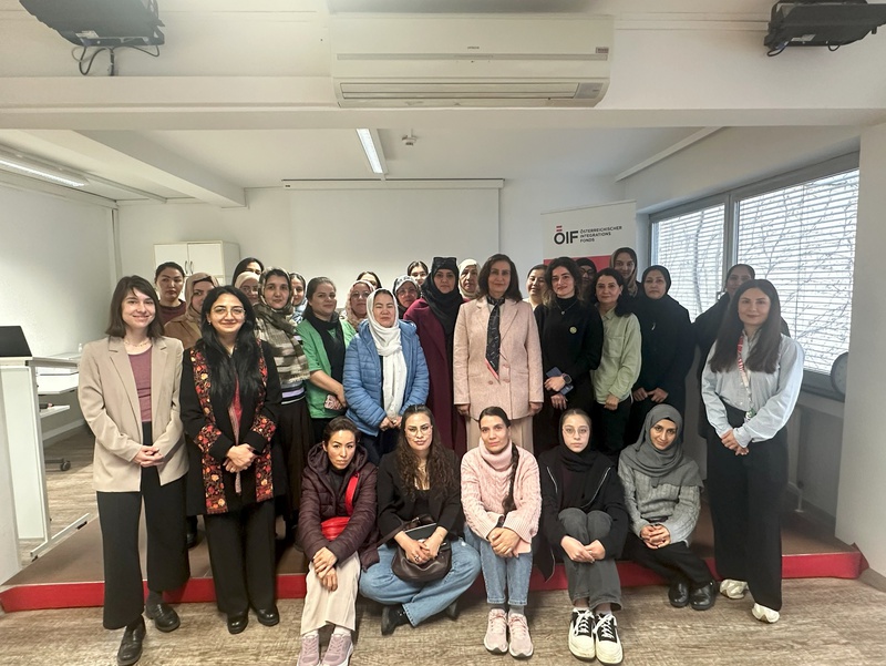 Gruppenfoto von etwa 30 Frauen in einem Raum. Die Teilnehmerinnen stehen auf einer roten Stufe und sind in unterschiedlichen Outfits gekleidet. Der Hintergrund zeigt Fenster und eine Präsentationswand mit einem Logo. Sie wirken konzentriert und freundlich.