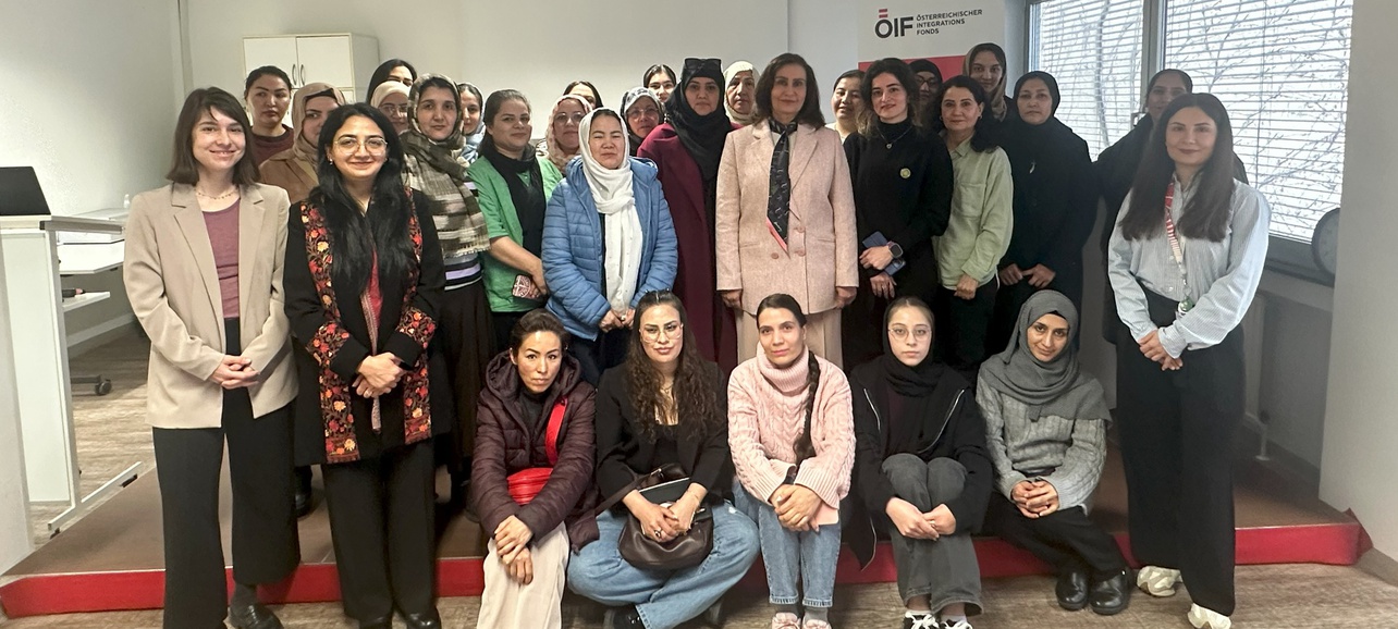 Gruppenfoto von etwa 30 Frauen in einem Raum. Die Teilnehmerinnen stehen auf einer roten Stufe und sind in unterschiedlichen Outfits gekleidet. Der Hintergrund zeigt Fenster und eine Präsentationswand mit einem Logo. Sie wirken konzentriert und freundlich.
