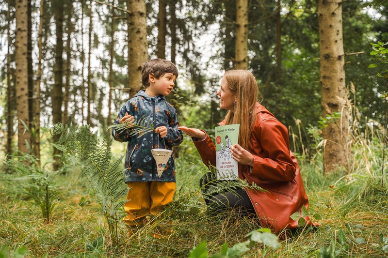Ein Kind und eine Erwachsene stehen im Wald, während sie sich über ein Lernmaterial austauschen.