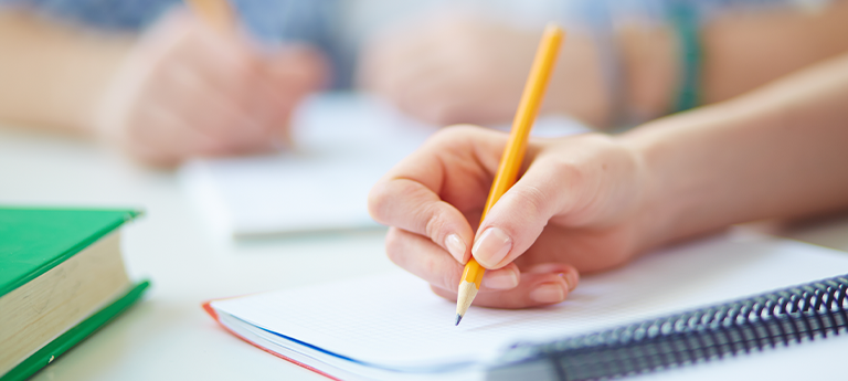 Eine Hand mit einem Bleistift schreibt auf ein leeres Notizbuch, während im Hintergrund weitere Hände sichtbar sind.