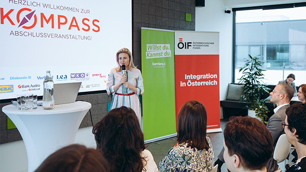Frau spricht bei einer Abschlussveranstaltung mit dem Titel "Kompass". Teilnehmer sind aufmerksam im Hintergrund.