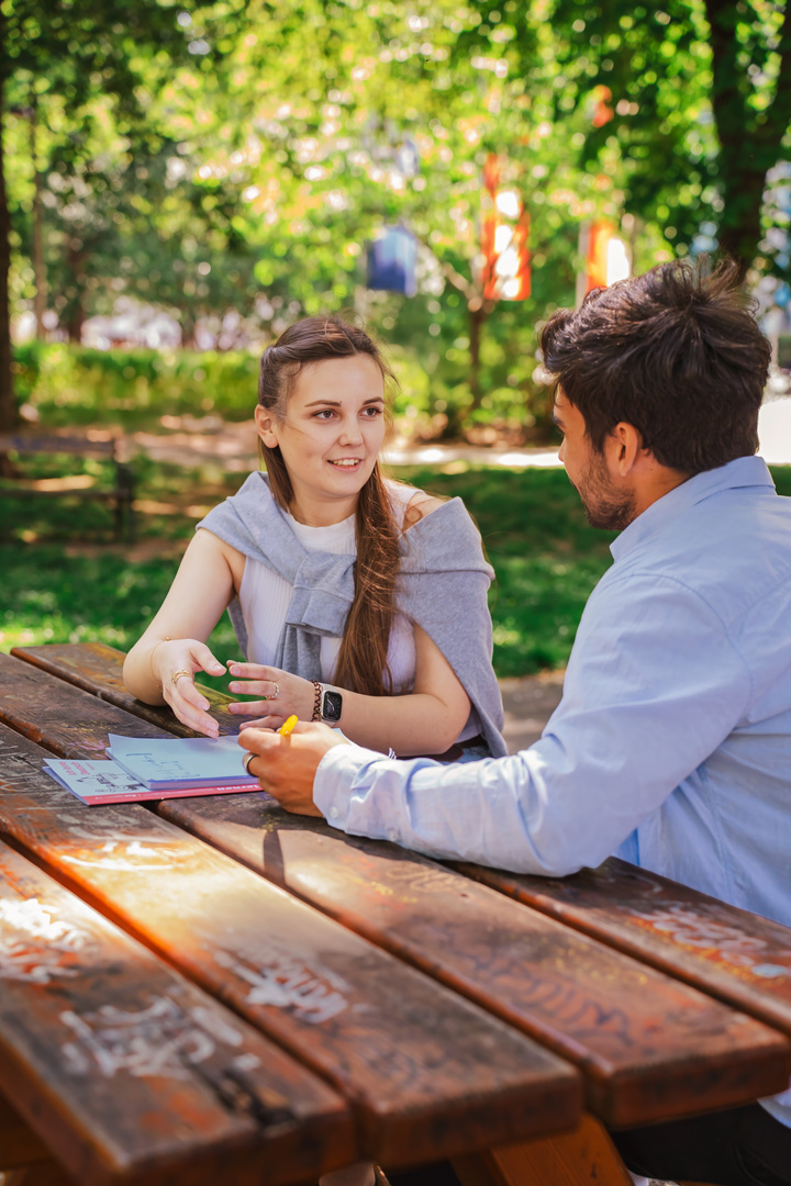 Zwei Personen sitzen an einem Tisch im Freien und unterhalten sich, während sie Notizen machen.