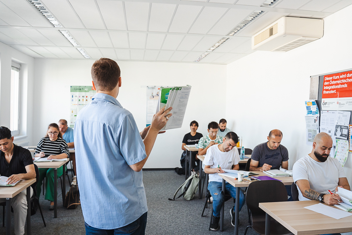 Trainer steht vor Klasse, Teilnehmer und Teilnehmerin schreiben