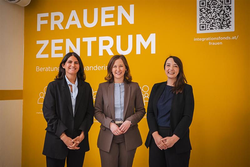 Drei Frauen stehen vor einer gelben Wand mit der Aufschrift "FRAUEN ZENTRUM". Sie tragen unterschiedliche formelle Kleidung und lächeln in die Kamera. Im Hintergrund sind Informationen über das Zentrum zu sehen.